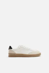 Zapatillas deportivas de estilo retro, color blanco con detalles en beige y negro. Capellada de cuero sintético con perforaciones laterales y puntera de gamuza. Suela de goma color caramelo.