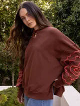 Buzo de algodón color blanco roto, de corte holgado, con puños y tajos laterales. Presenta un bordado artesanal en relieve de líneas orgánicas en color marrón caramelo sobre las mangas.