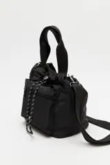 Cartera tipo bolso de nylon negro, con diseño utilitario y formato flexible que se ajusta con cordón en la parte superior. Posee asas de mano y una correa ajustable y desmontable para llevar cruzada. Incluye bolsillo frontal con cierre y herrajes metálicos.