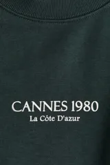 Buzo de tejido de punto color verde oscuro, con cuello redondo, hombros caídos y puños y bajo acanalados. Presenta un pequeño estampado centrado en el pecho con el texto "CANNES 1980" y debajo "La Côte D'Azur".