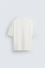 Camiseta blanca de corte holgado, cuello redondo y mangas cortas. Presenta un bordado y estampado combinado en el frente.