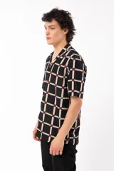Camisa de manga corta con calce relajado y cuello tipo sastre, confeccionada en rayón. Presenta un diseño de cuadrícula beige sobre fondo negro con detalles en azul y rojo en las intersecciones.
