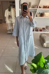 Vestido camisero color celeste de lino, con cuello clásico, cierre frontal con botones y mangas 3/4. Tiene dos bolsillos de parche en el pecho y corte irregular en el ruedo.