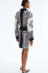 Vestido camisero de manga larga, con cuello camisero y cierre frontal de botones ocultos. Presenta un estampado tipo pañuelo en tonos blanco, azul marino y azul claro, y se ajusta a la cintura con un cinturón extraíble con argollas metálicas.