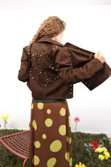 Chaqueta marrón de algodón con botones metálicos, cuello clásico, bolsillos frontales y apliques de tachas y botones formando una flor en la espalda.