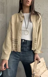 Sobrecamisa beige de gamuza con cuello camisero, cierre frontal con botones y mangas largas con puños abotonados. Presenta bolsillos de parche en el pecho y detalle de calado en el ruedo.