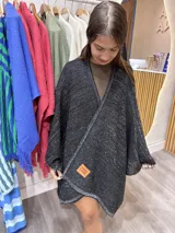 Ruana tejida de punto color negro con hilos de lurex que le dan un brillo sutil. Diseño envolvente con cuello en V y bordeado en hilo plateado. Posee una etiqueta de cuero marrón con el texto "PURA LANA" en el frente.