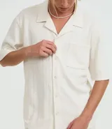 Camisa de jacquard color crema, de corte oversized, con cuello solapa, manga corta, abotonadura frontal, bolsillo en el pecho y textura en relieve.