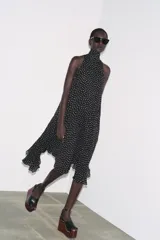 Vestido midi negro con estampado de lunares blancos, cuello halter con pañuelo integrado, sin mangas y bajo asimétrico. Cierre con abertura y botón en la nuca.
