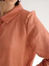 Camisa blanca de lino con cuello clásico, abotonada al frente con botones de madera, mangas largas con volados en los hombros y corte femenino.