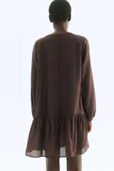 Vestido corto marrón oscuro, estilo túnica, con escote redondo amplio y botones forrados en el frente. Presenta mangas globo largas estilo raglán con puños elásticos y un fruncido escalonado bajo la cadera que le da volumen.