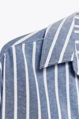Camisa de algodón a rayas verticales azules y blancas con hilo metalizado, cuello solapa y escote pico. Manga larga acabada en puño, bajo asimétrico y cierre frontal con botones.