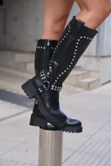 Bota alta de caña ancha color negro, estilo biker o motera, con suela track gruesa y taco bajo. Presenta apliques de tachas plateadas a lo largo de la caña y una correa con hebilla y argolla decorativa alrededor del tobillo.