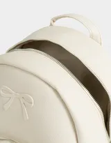Mochila pequeña de cuerina color rosa pálido, con moño en relieve en el frente y llavero con forma de corazón trenzado. Tiene correas regulables, manija superior, cierre superior, bolsillo frontal con cierre y bolsillos laterales.