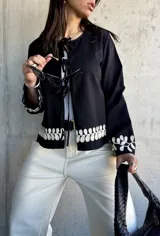 Chaqueta negra con escote redondo y cierre frontal con lazos. Presenta bordado de hojas blancas en los puños.