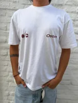 Remera blanca de corte oversize con cuello redondo y manga corta. Presenta un estampado frontal en color marrón rojizo con un diseño abstracto que incluye un sol estilizado y la palabra "Osten".
