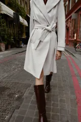 Bota alta de cuero sintético color marrón oscuro, con caña recta y punta cuadrada. Presenta pespuntes a contraste en las uniones y un detalle de correa con hebilla metálica en la parte superior. Posee un tacón de bloque de altura media.