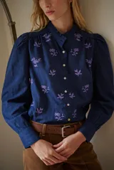 Camisa azul marino con estampado floral en color lila, cuello mao con volantes, cierre frontal de botones y mangas largas abullonadas con puños elásticos.