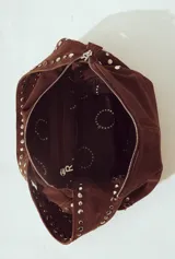 Bolso de hombro tipo hobo, color camel, confeccionado en gamuza o material similar con acabado afelpado. Presenta detalles de tachas metálicas plateadas a lo largo de los bordes laterales y en la correa de hombro.