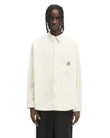 Camisa de manga larga confeccionada en pana de color blanco roto, con cuello clásico y cierre frontal de botones. Presenta un bolsillo de parche en el pecho con la etiqueta de la marca Carhartt.
