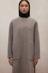 Abrigo largo de lana reciclada color gris topo, con cuello redondo y cierre frontal de botones ocultos. Presenta bolsillos amplios y rectangulares en la parte delantera.