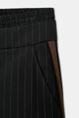 Pantalón negro con finas rayas verticales blancas, de tiro medio y corte ancho. Presenta bolsillos delanteros y traseros, cintura doble tipo bóxer con botones y cierre frontal con cremallera y ganchos metálicos. Detalle de franja marrón en los laterales.