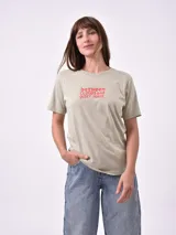 Remera blanca de algodón con cuello redondo y manga corta. Presenta un pequeño bordado en el pecho izquierdo con una figura que parece ser una persona acostada en una tabla de surf o flotador.