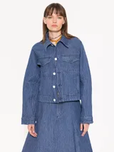 Campera de denim azul con textura en relieve, corte recto, cuello camisero, cierre frontal con botones blancos, bolsillos delanteros y mangas largas con pinzas que generan volumen.