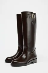 Bota alta de cuero color marrón oscuro, con caña alta, punta redonda y hebilla decorativa en la parte superior.