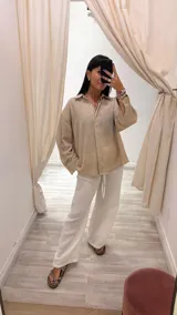 Camisa color beige claro con textura brillante, cuello clásico, cierre frontal con botones y mangas largas con puños abotonados.