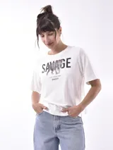 Remera blanca de manga corta con cuello redondo y estampado frontal que dice "SAVAGE" con una figura de animal (posiblemente un leopardo o jaguar) hecho de lentejuelas o material brillante. Debajo del estampado principal se lee "LIFE GOES ON ENJOY".