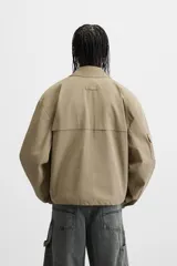 Cazadora color arena de corte holgado, con cuello solapa y manga larga con bolsillo multifunción. Tiene bolsillos frontales de plastrón, bolsillo interior y elástico en los puños y ruedo. El ruedo es ajustable con cordón. Cierre frontal con cremallera oculta por solapa con botones a presión.