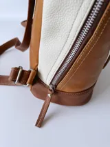 Mochila pequeña de cuero color camel, con base marrón, laterales color crema y cierre superior con cremallera.