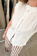 Blusa blanca sin mangas con cuello mao, volados en cuello, sisas y ruedo, y cierre frontal con botones.