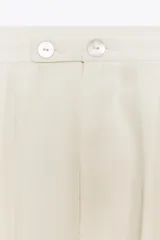 Pantalón palazzo color crudo, de tiro alto, confeccionado en lyocell. Presenta bolsillos laterales ocultos, detalle de pliegues en el delantero y cierre frontal con cremallera y botones.