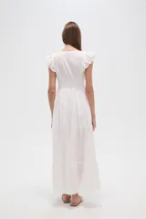 Vestido largo blanco de algodón con escote en V, manga corta con volados de broderie y talle entallado con botones frontales. La falda es amplia y presenta detalles de bordados calados en el ruedo.