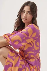 Vestido midi cruzado con estampado floral abstracto en tonos violeta y naranja, con mangas 3/4 abullonadas y lazo ajustable en la cintura. Presenta un tajo lateral.