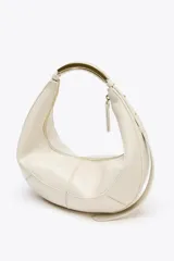 Bolso ovalado color crudo con asa de hombro metálica dorada. Tiene pespuntes en contraste, bolsillo interior con cremallera y cierre principal con cremallera y tirador.