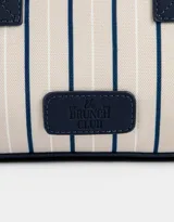 Cartera bowling con estampado de rayas verticales color beige y azul marino. Tiene doble asa corta de mano y correa regulable y desmontable. Incluye cierre superior y tag frontal de cuerina con el logo 'The Brunch Club'.