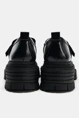 Zapato tipo mocasín color negro, con plataforma y suela track. Presenta una tira ajustable adherente sobre el empeine y tirador trasero.