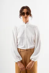 Camisa blanca de manga larga con cuello alto fruncido y puños ajustados. Presenta un corte holgado y tela con textura ligera.