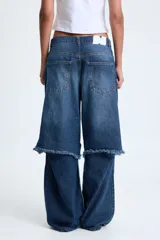 Jean tiro bajo de denim rígido 100% algodón, color azul índigo profundo con matices grises. Presenta un calce recto con pierna amplia y un diseño innovador con corte a la altura de la rodilla, creando un efecto de dos piezas superpuestas con terminación deshilachada.
