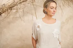 Blusa color crema de lino con escote redondo y mangas cortas. Presenta apliques florales de razo y prendedor de strass en el frente.
