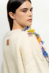 Sweater color crema de tejido de punto con cuello redondo y mangas largas. Presenta flecos de colores en los hombros.