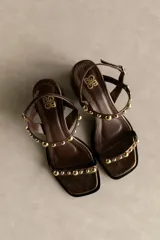 Sandalia de cuero color marrón con taco bloque medio y punta cuadrada. Presenta tachas esféricas doradas en las tiras y hebilla de ajuste en el tobillo.