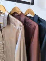 Camisa marrón de satén con cuello clásico, cierre frontal con botones y mangas largas con puños abotonados.