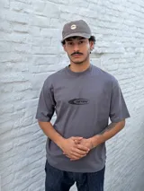 Remera gris de corte oversize con cuello redondo y manga corta. Estampado frontal con el texto "OSTEN" en negro con un diseño estilizado.