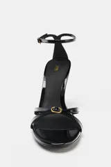 Sandalia negra acharolada de tacón fino alto, con tiras delanteras y cierre de pulsera al tobillo con hebilla.