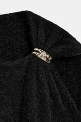 Jersey de punto negro, sin mangas (manga sisa), con cuello cuadrado drapeado y un broche dorado decorativo en el hombro.