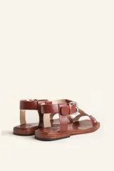 Sandalia de cuero color marrón con tiras finas, detalles de tachas metálicas y hebilla lateral.